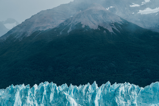 View of the Perito Moreno Glacier with its jagged blue ice formations set against a backdrop of dark forested mountains in Perito Moreno Santa Cruz Province Argentina.
