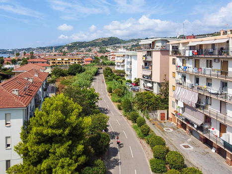 Aerial view of a paved cycle path lined with green trees and multi story residential buildings under a bright blue sky with distant hills in San Lorenzo Liguria Italy.