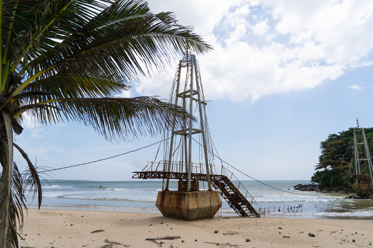 View of a metal bridge structure on the sandy beach with palm tree leaves and ocean waves under a cloudy sky in Matara Southern Province Sri Lanka.