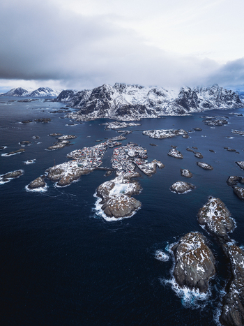 Aerial view of the rugged snow dusted islands rise from the dark swirling sea a stark contrast against the distant snow capped mountains Henningsv&aelig;r Nordland Norway.