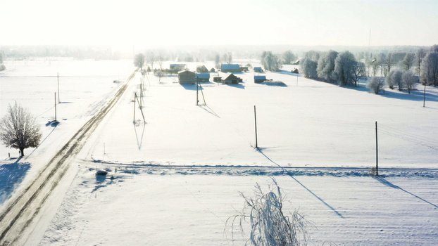 Aerial view of a snow covered rural landscape with scattered farm buildings frost covered trees and a quiet road under bright winter sunlight in Latvia. Aerial view of a snow covered rural landscape with scattered farm buildings frost covered trees and a quiet road under bright winter sunlight in Latvia.