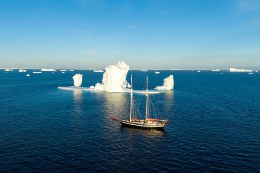 Aerial view of a sailboat navigating near a large arch iceberg in the deep blue waters of Greenland. Aerial view of a sailboat navigating near a large arch iceberg in the deep blue waters of Greenland.