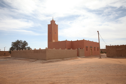 View of a traditional earthen mosque with a minaret situated in an arid desert landscape under a clear blue sky in Morocco.