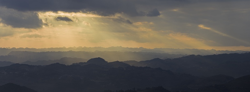 Aerial view of rolling mountain ridges and silhouetted hills under a dramatic sunset sky with golden sunbeams breaking through dark clouds in Gaoxing Miao Village Guizhou China. Aerial view of rolling mountain ridges and silhouetted hills under a dramatic sunset sky with golden sunbeams breaking through dark clouds in Gaoxing Miao Village Guizhou China.