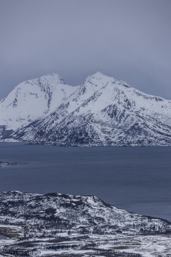 View of snow capped mountain peaks rising above a calm fjord under an overcast winter sky Kvaloya Troms Norway.