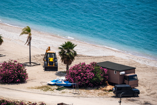 View of a yellow excavator on a pebble beach with palm trees pink oleander bushes and a small cabin by the turquoise sea in Vlore County Albania. View of a yellow excavator on a pebble beach with palm trees pink oleander bushes and a small cabin by the turquoise sea in Vlore County Albania.
