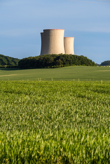 View of nuclear power plant cooling towers rising above a lush green field and forest in Czechia. View of nuclear power plant cooling towers rising above a lush green field and forest in Czechia.