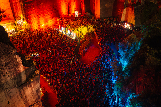 Aerial view of a large crowd of people attending an outdoor music concert inside a deep stone quarry illuminated with vibrant red and blue stage lighting in Vintijan Istria County Croatia. Aerial view of a large crowd of people attending an outdoor music concert inside a deep stone quarry illuminated with vibrant red and blue stage lighting in Vintijan Istria County Croatia.