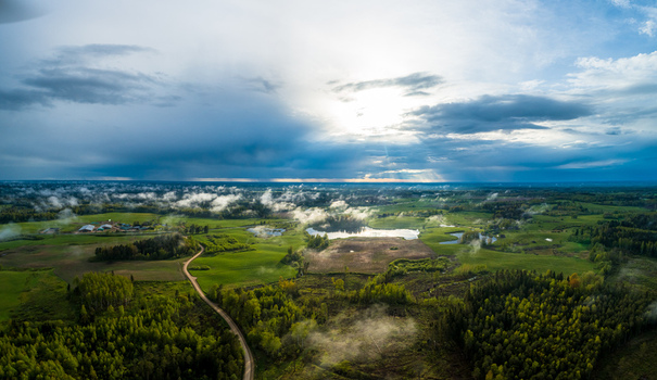 Aerial view of a rural landscape featuring lush green fields dense forests a small lake and a winding dirt road under a dramatic sky with sunbeams breaking through clouds Latvia. Aerial view of a rural landscape featuring lush green fields dense forests a small lake and a winding dirt road under a dramatic sky with sunbeams breaking through clouds Latvia.