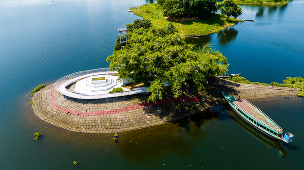 Aerial view of a small island in Kaptai Lake featuring a white monument a large tree and a traditional wooden boat moored at the stone paved shore in Rangamati Chittagong Division Bangladesh.