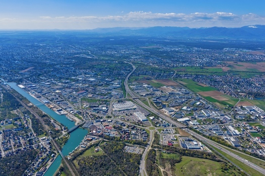 Aerial view of a sprawling cityscape highways cutting through the urban fabric a canal reflecting the sky and distant mountains Sausheim Grand Est France.