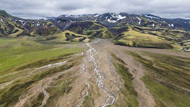 Aerial view of a braided river system winding through the mossy green highlands with snow capped mountains under a cloudy sky in Iceland. Aerial view of a braided river system winding through the mossy green highlands with snow capped mountains under a cloudy sky in Iceland.
