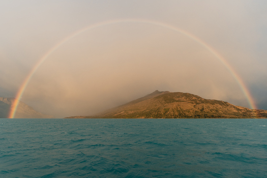 View of a vibrant rainbow arching over the turquoise waters of Lago Argentino with a mountain landscape in the background in El Calafate Santa Cruz Province Argentina.