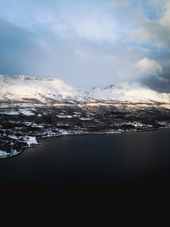 Aerial view of snow capped mountains meet the dark still waters of the fjord contrasting sharply under a brooding sky Sjøvegan Troms Norway. Aerial view of snow capped mountains meet the dark still waters of the fjord contrasting sharply under a brooding sky Sjøvegan Troms Norway.