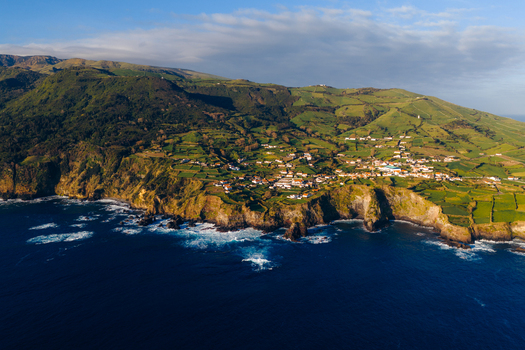Aerial view of a coastal village on rugged cliffs overlooking the Atlantic Ocean with lush green fields and the Farol de Albarnaz lighthouse in Ponta Delgada Acores Portugal. Aerial view of a coastal village on rugged cliffs overlooking the Atlantic Ocean with lush green fields and the Farol de Albarnaz lighthouse in Ponta Delgada Acores Portugal.