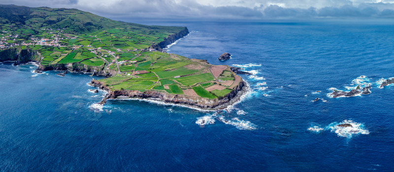 Aerial view of vibrant green fields meet the rugged coastline where the Estrada do Farol winds along the cliffs Flores A&ccedil;ores Portugal.