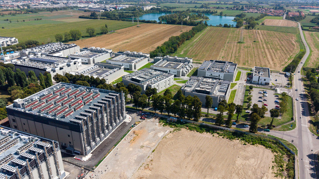 Aerial view of a large data center campus featuring multiple industrial buildings with rooftop cooling systems parking areas and surrounding green fields in Milan Lombardy Italy. Aerial view of a large data center campus featuring multiple industrial buildings with rooftop cooling systems parking areas and surrounding green fields in Milan Lombardy Italy.