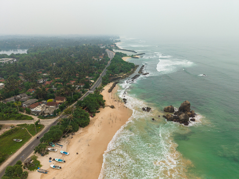 Aerial view of the sandy coastline with turquoise waves crashing against a large rock formation and a coastal road lined with lush palm trees in Koggala Southern Province Sri Lanka. Aerial view of the sandy coastline with turquoise waves crashing against a large rock formation and a coastal road lined with lush palm trees in Koggala Southern Province Sri Lanka.