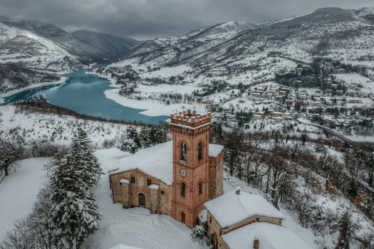 Aerial view of a historic brick church covered in snow situated on a hill overlooking a turquoise lake and a snow covered village in Fiastra Marche Italy. Aerial view of a historic brick church covered in snow situated on a hill overlooking a turquoise lake and a snow covered village in Fiastra Marche Italy.