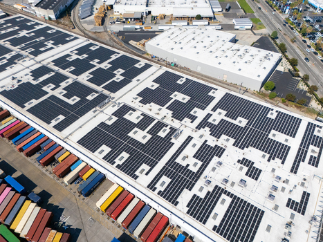 Aerial view of a large industrial warehouse facility featuring extensive rooftop solar panel arrays and a yard filled with colorful shipping containers in Carson California United States.