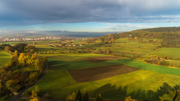 Aerial view of rolling green fields and agricultural land with autumn trees and a distant town under a cloudy sky in Germany. Aerial view of rolling green fields and agricultural land with autumn trees and a distant town under a cloudy sky in Germany.