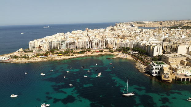 Aerial view of the coastal urban landscape with dense residential buildings a rocky shoreline and numerous boats anchored in the turquoise Mediterranean Sea in Malta. Aerial view of the coastal urban landscape with dense residential buildings a rocky shoreline and numerous boats anchored in the turquoise Mediterranean Sea in Malta.