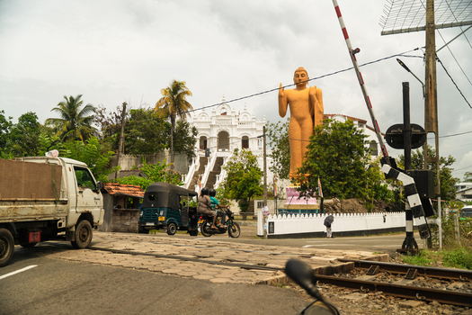 View of a large golden Buddha statue and a white temple near a railway crossing with local traffic in Dodanduwa Southern Province Sri Lanka. View of a large golden Buddha statue and a white temple near a railway crossing with local traffic in Dodanduwa Southern Province Sri Lanka.