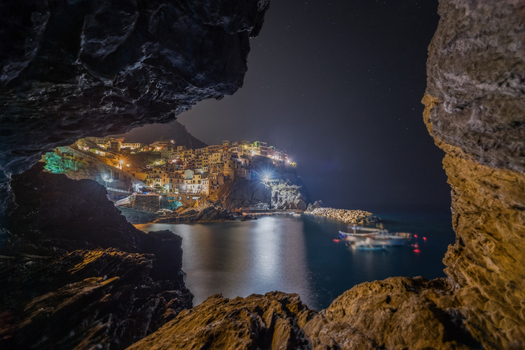View of the illuminated coastal village of Manarola at night seen through a rocky cave opening with boats in the harbor and lights reflecting on the water Manarola Liguria Italy. View of the illuminated coastal village of Manarola at night seen through a rocky cave opening with boats in the harbor and lights reflecting on the water Manarola Liguria Italy.