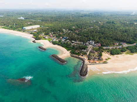 Aerial view of turquoise ocean waves hitting sandy beaches and stone breakwaters near a coastal village surrounded by lush palm forests Peraliya Southern Province Sri Lanka. Aerial view of turquoise ocean waves hitting sandy beaches and stone breakwaters near a coastal village surrounded by lush palm forests Peraliya Southern Province Sri Lanka.