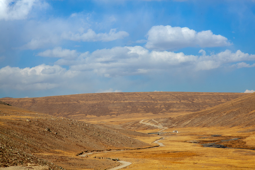 View of the vast arid landscape of Deosai National Park with a winding dirt road cutting through rolling hills under a bright blue sky in Astore Gilgit Pakistan. View of the vast arid landscape of Deosai National Park with a winding dirt road cutting through rolling hills under a bright blue sky in Astore Gilgit Pakistan.