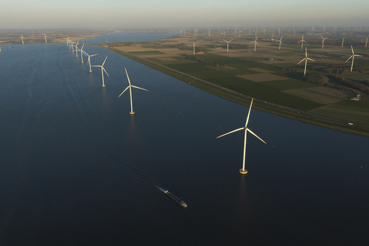 Aerial view of modern wind turbines standing in the water and on green fields along a canal with a cargo barge at sunset in the Netherlands. Aerial view of modern wind turbines standing in the water and on green fields along a canal with a cargo barge at sunset in the Netherlands.