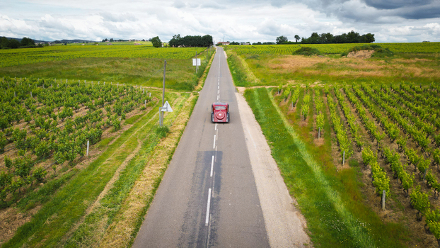Aerial view of a vintage red car driving on a straight road through lush green vineyards under a cloudy sky La Chapelle de Guinchay Bourgogne Franche Comte France.