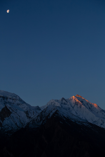 View of the snow capped peaks of Rakaposhi mountain with golden sunlight on the summit and a crescent moon in the deep blue twilight sky Hunza Nagar Gilgit Pakistan. View of the snow capped peaks of Rakaposhi mountain with golden sunlight on the summit and a crescent moon in the deep blue twilight sky Hunza Nagar Gilgit Pakistan.