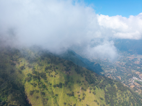 Aerial view of Wonokerto mountainside with lush green vegetation scattered trees and rolling hills partially obscured by soft white clouds in Sukorejo East Java Indonesia. Aerial view of Wonokerto mountainside with lush green vegetation scattered trees and rolling hills partially obscured by soft white clouds in Sukorejo East Java Indonesia.