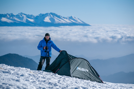 Liptovsky Jan Zilina Region Slovakia 09 March 2025 View of a man in a blue jacket standing by a green tent on the snow covered peak of Dumbier with clouds and distant mountains. Liptovsky Jan Zilina Region Slovakia 09 March 2025 View of a man in a blue jacket standing by a green tent on the snow covered peak of Dumbier with clouds and distant mountains.