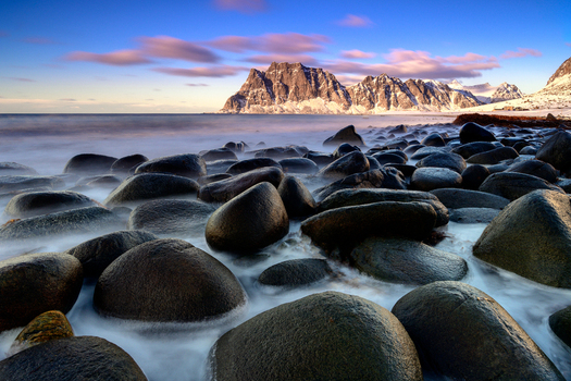 View of smooth dark boulders on a sandy beach with snow capped mountains under a golden sunset sky in Leknes Lofoten Norway.