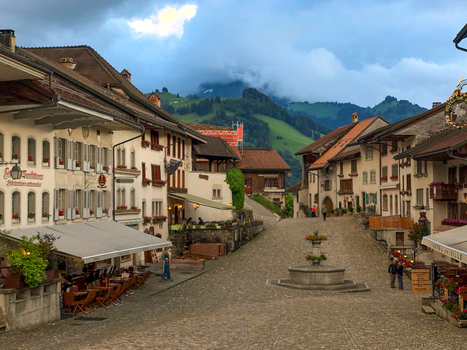 Gruyeres Switzerland 14 May 2026 View of the historic cobblestone street with traditional buildings a stone fountain and lush green hills under a cloudy sky. Gruyeres Switzerland 14 May 2026 View of the historic cobblestone street with traditional buildings a stone fountain and lush green hills under a cloudy sky.