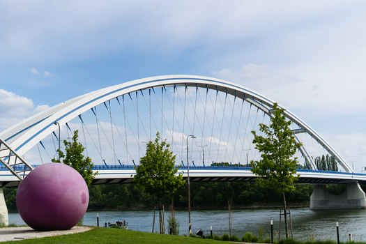 View of the Apollo Bridge with its white arch over the Danube river and a large purple sphere sculpture on the grassy bank in Bratislava Bratislava Region Slovakia. View of the Apollo Bridge with its white arch over the Danube river and a large purple sphere sculpture on the grassy bank in Bratislava Bratislava Region Slovakia.