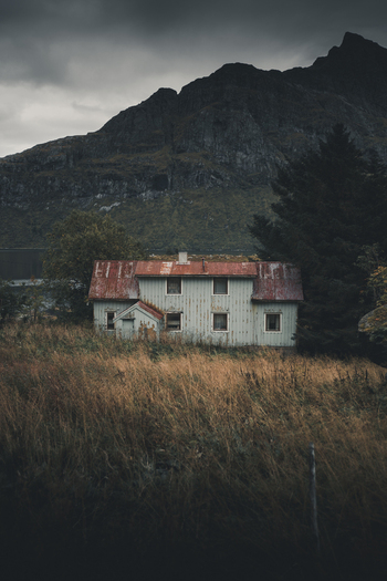 Nordland Norway 09 September 2022 View of a weathered wooden house with a red roof set in a field of tall dry grass against a backdrop of dramatic dark mountains in the Lofoten islands. Nordland Norway 09 September 2022 View of a weathered wooden house with a red roof set in a field of tall dry grass against a backdrop of dramatic dark mountains in the Lofoten islands.