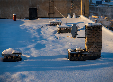 View of a snow covered rooftop featuring a brick chimney a satellite dish and ventilation units under soft winter sunlight in Vilnius Vilnius County Lithuania. View of a snow covered rooftop featuring a brick chimney a satellite dish and ventilation units under soft winter sunlight in Vilnius Vilnius County Lithuania.