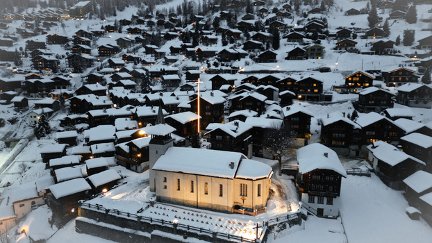Aerial view of a snow covered village with a brightly lit church standing prominently amidst the dark rustic chalets Grimentz Valais Switzerland.