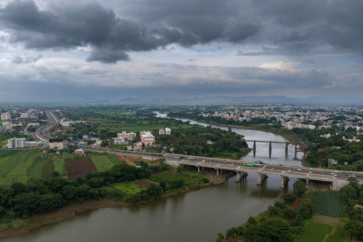 Aerial view of the Krishna River flowing through the city of Karad with bridges lush green agricultural fields and urban buildings under a cloudy sky in Karad Maharashtra India.