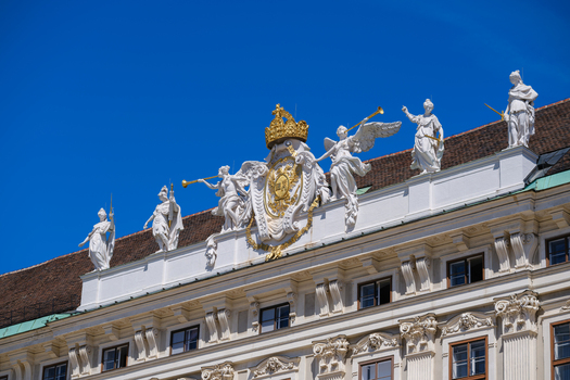 View of the Hofburg palace roofline featuring white statues and a golden coat of arms under a clear blue sky in Vienna Vienna Austria. View of the Hofburg palace roofline featuring white statues and a golden coat of arms under a clear blue sky in Vienna Vienna Austria.