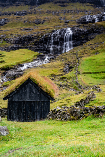 View of a traditional black wooden hut with a grass roof situated in a lush green landscape with a waterfall cascading down a rocky hillside in the Faroe Islands.