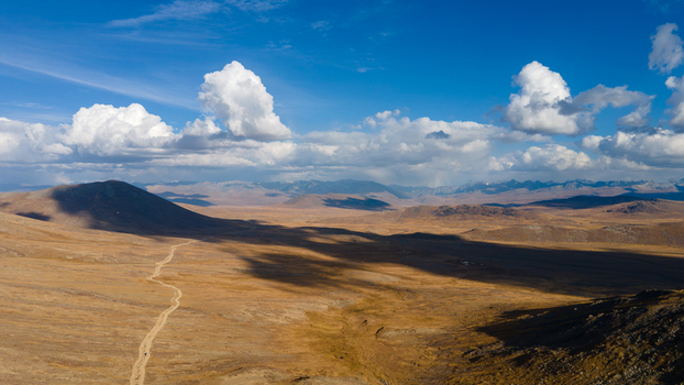 Aerial view of Deosai Plains with a winding dirt road cutting through the vast plateau surrounded by distant mountains under a blue sky with clouds in Skardu Gilgit Pakistan.