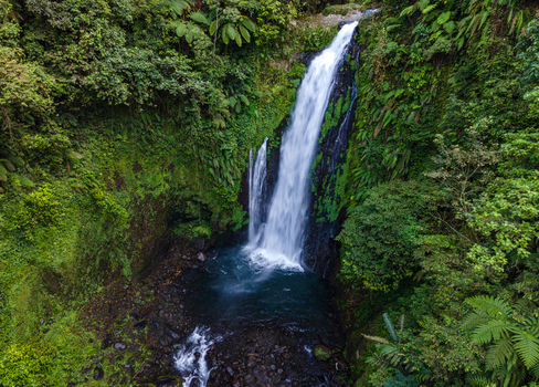 Aerial view of Gomblang Waterfall cascading into a dark pool surrounded by dense tropical jungle and lush green foliage in Kecamatan Kedungbanteng Jawa Tengah Indonesia. Aerial view of Gomblang Waterfall cascading into a dark pool surrounded by dense tropical jungle and lush green foliage in Kecamatan Kedungbanteng Jawa Tengah Indonesia.