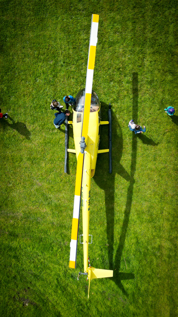 Aerial view of a yellow helicopter parked on a green grass field with people standing nearby and long shadows in La Chapelle de Guinchay Bourgogne Franche Comte France.