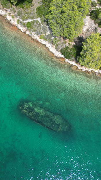 Aerial view of a submerged shipwreck in the clear turquoise waters near the rocky coastline and green pine trees in Necujam Split Dalmatia County Croatia. Aerial view of a submerged shipwreck in the clear turquoise waters near the rocky coastline and green pine trees in Necujam Split Dalmatia County Croatia.
