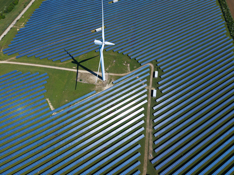 Aerial view of Chelveston Wind Farm featuring a tall wind turbine casting a long shadow over rows of blue solar panels on green fields in Wellingborough England United Kingdom. Aerial view of Chelveston Wind Farm featuring a tall wind turbine casting a long shadow over rows of blue solar panels on green fields in Wellingborough England United Kingdom.