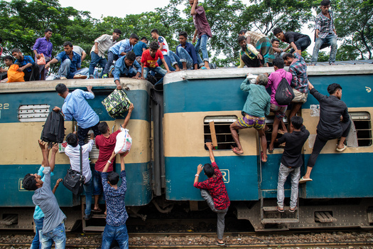 Dhaka Bangladesh 14 June 2018 View of people climbing and sitting on the roof of a crowded blue and yellow train at Airport Railway Station with lush green trees in the background. Dhaka Bangladesh 14 June 2018 View of people climbing and sitting on the roof of a crowded blue and yellow train at Airport Railway Station with lush green trees in the background.